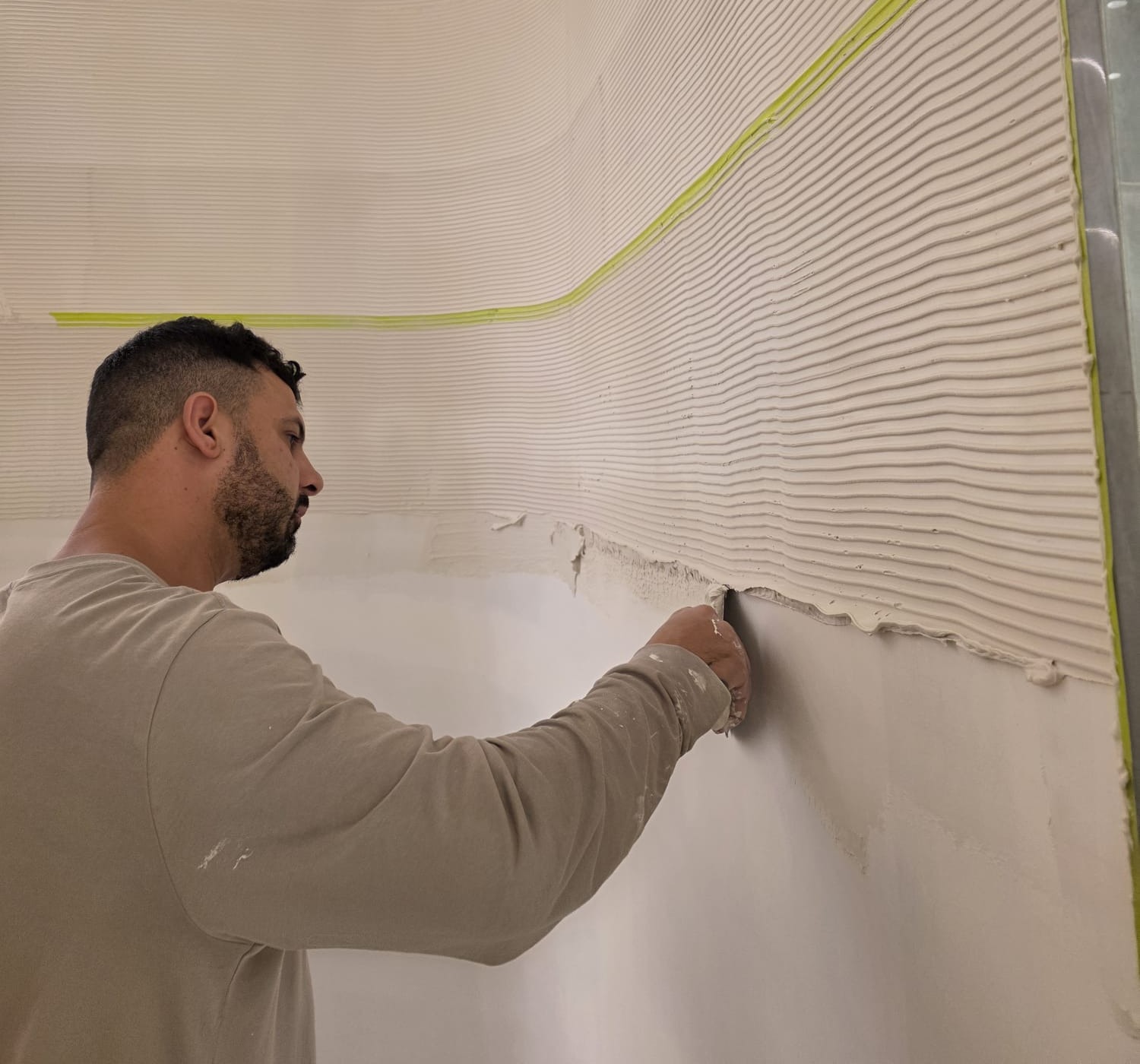 Giovanni hand-applying a combed, textured plaster finish to a wall, with masking tape lines marking the transition between sections.